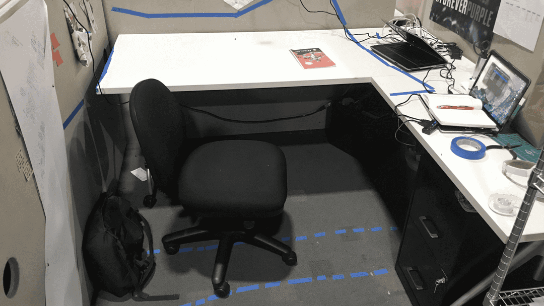 Small cubicle with a laptop pointed towards one corner, and areas denoted with blue tape